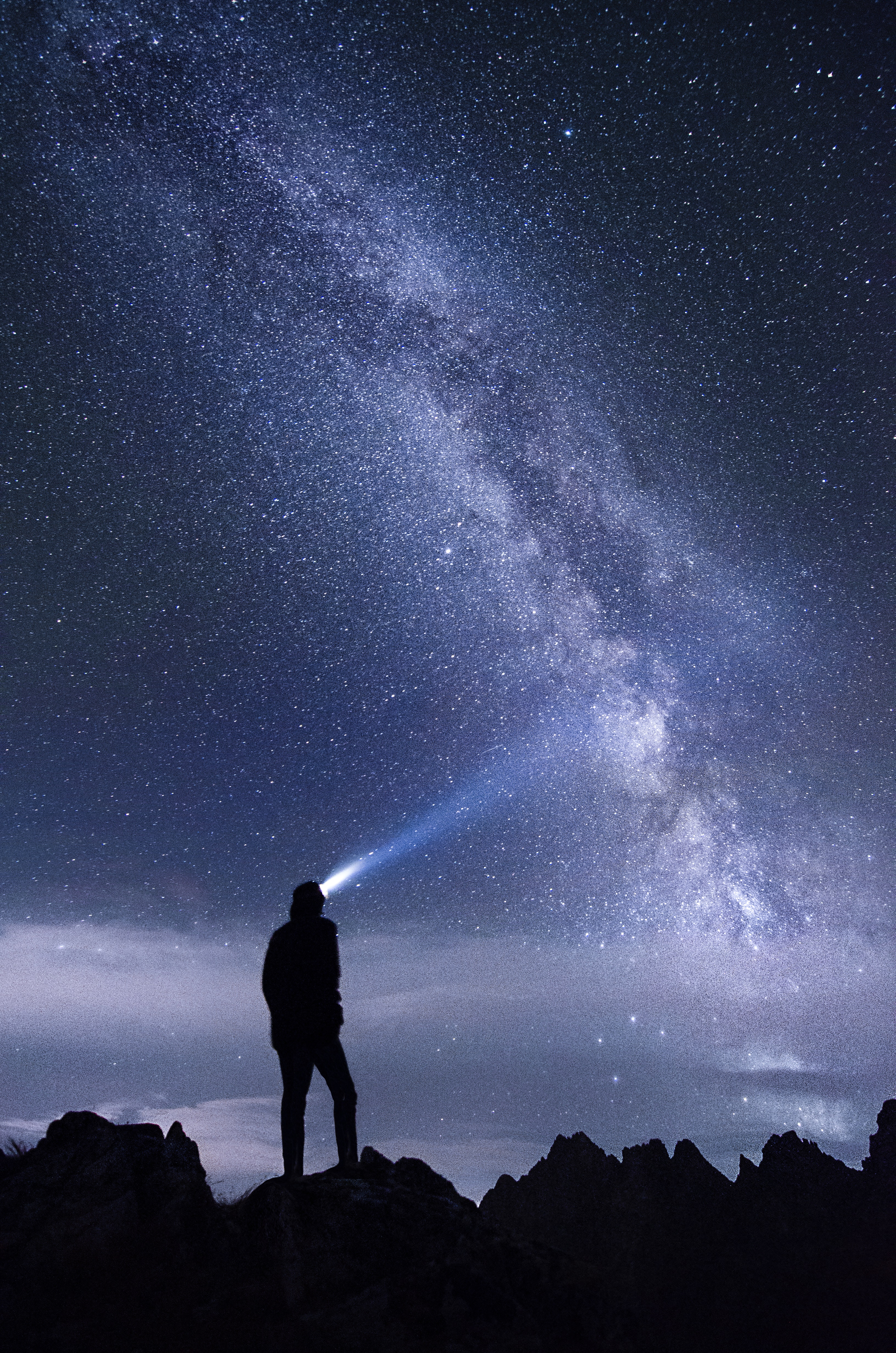 A person is silhouetted standing on top of a jagged ridge with their headlamp shining skyward. The Milky Way blazes bright across a star filled sky.